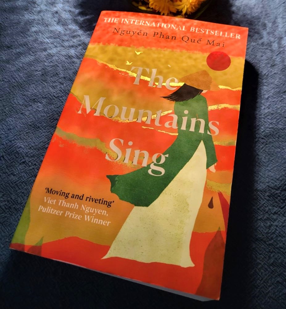 The Book Cover of The Mountains Sing. It is held in warm golden-orange tones and shows a very minimal painted figurine with long black hair, a green coat, a white dress and a typical vietnamese hat. 